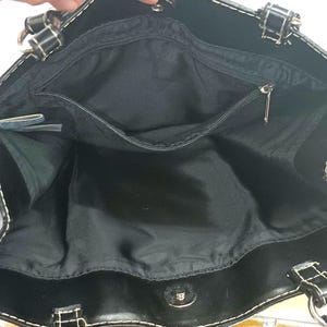 May include: Black tote bag with a spacious interior, featuring a zippered pocket and a magnetic snap closure. The bag has white stitching and silver-tone hardware. The handles are black with silver rings.