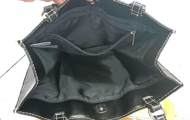May include: Black tote bag with a spacious interior, featuring a zippered pocket and a magnetic snap closure. The bag has white stitching and silver-tone hardware. The handles are black with silver rings.