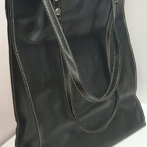 May include: Black tote bag with white stitching and silver hardware. The bag has two handles and a shoulder strap. The bag is made of a smooth material and has a rectangular shape. The bag is sitting on a white surface.
