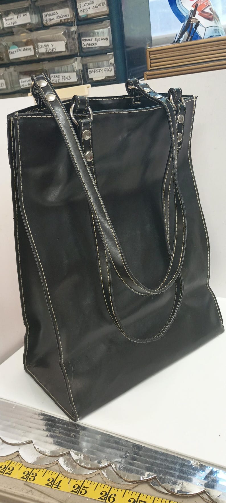 May include: Black tote bag with white stitching and silver hardware. The bag has two handles and a shoulder strap. The bag is made of a smooth material and has a rectangular shape. The bag is sitting on a white surface.