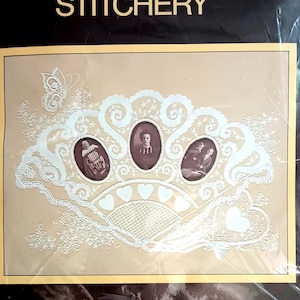May include: Sunset Stitchery kit featuring a Victorian Photo Gallery design. The kit includes a cream-colored fan-shaped design with three oval portraits and delicate white lace details. The design fits a 14" x 18" frame.