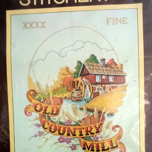 May include: Sunset Stitchery kit featuring an embroidered scene of an old country mill. The design includes a red mill building, water, trees, and the words "Old Country Mill" and "All Grains". The frame size is 14" x 18".