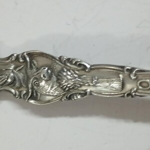 May include: A detailed close-up of a silver spoon with ornate carvings. The spoon's handle features a classical face, a bird, and text. The spoon is made of a shiny, reflective metal, likely silver.