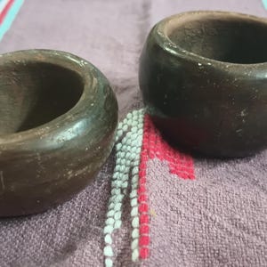 May include: Two handcrafted, dark brown wooden napkin rings. The rings have a smooth, polished finish and a rounded shape. They are displayed on a purple cloth with colorful striped accents. These napkin rings are a rustic addition to any table setting.