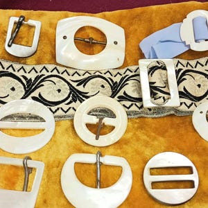 May include: Assortment of vintage white mother of pearl belt buckles with metal clasps. The buckles are arranged on a yellow fabric background.