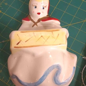 May include: A vintage ceramic figurine of a woman in a bonnet and red cape, holding a yellow object with brown accents. The figurine has a white base with a blue wave design. The woman has blonde hair, blue eyes, and red lips.