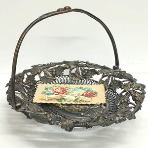 May include: A silver-toned wire basket with a handle, featuring a decorative floral design. The basket is empty, but there is a vintage greeting card with a floral design inside.
