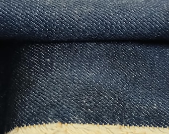 May include: Close-up of a folded piece of dark blue denim fabric. The fabric has a textured weave with small white flecks throughout. The edge of the fabric is frayed, revealing a beige fringe.