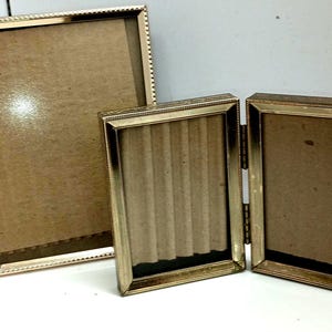 May include: Two gold-toned picture frames. The larger frame is rectangular and has a beaded border. The smaller frame is a tri-fold design with three rectangular openings.