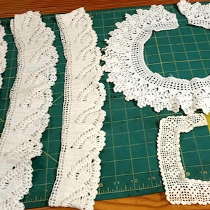 May include: Several pieces of white crochet lace are arranged on a green cutting mat. The pieces include long strips with a leaf-like pattern, a circular collar, and a square frame, all in a delicate, openwork design. The lace is likely for crafting or embellishment.