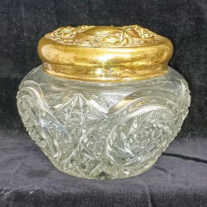 May include: A clear glass jar with a cut glass design and a gold metal lid. The lid has a decorative floral design.