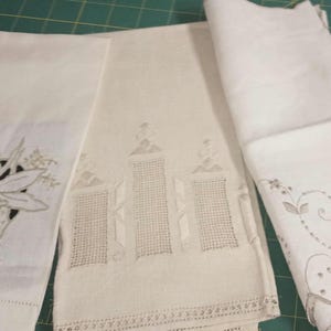 May include: Three white linen towels with embroidered designs. One towel features a floral design with black and gray accents. Another has a geometric pattern, and the third has a floral and leaf design with gray accents. The towels are folded and arranged on a green surface.