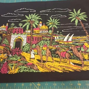 May include: Black fabric with a colorful illustration of a Middle Eastern scene. The image depicts camels, palm trees, buildings, and figures in traditional attire. The artwork has a vintage, travel-themed aesthetic.