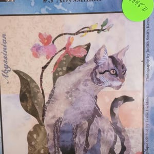 May include: A quilt pattern for a cat with a blue and gray patterned design, sitting in front of a pink and yellow flower. The pattern is titled "My Cats Garden" and is part of a "Block of the Month Quilt" series. The pattern is number 5 and is titled "Abyssinian".