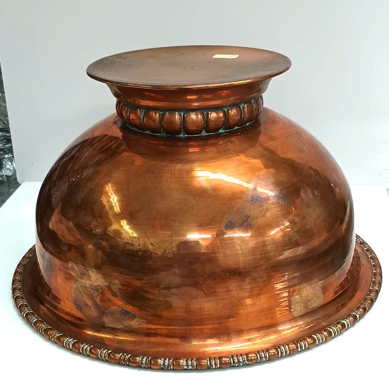 May include: A large copper bowl with a raised rim and a beaded design around the top and bottom. The bowl is polished and has a shiny finish.