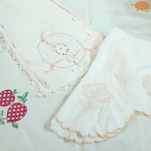 May include: White vintage linens with embroidered floral and fruit designs. The linens include a rectangular piece with a floral center and lace trim, and a scalloped-edge piece. The designs feature pink, green, and blue accents.