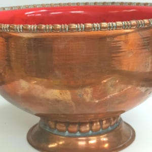 May include: A large copper bowl with a red interior and a decorative beaded rim. The bowl has a wide base and a rounded shape.