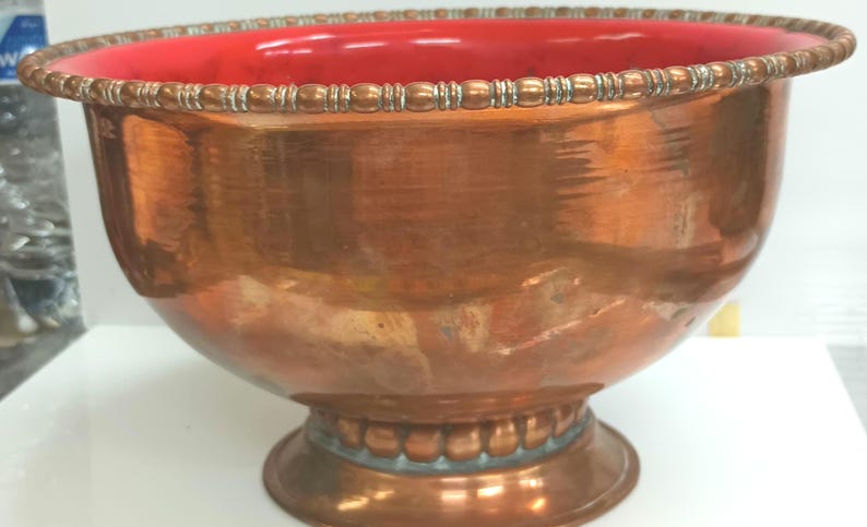 May include: A large copper bowl with a red interior and a decorative beaded rim. The bowl has a wide base and a rounded shape.