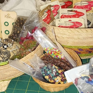 May include: A collection of craft supplies, including buttons, beads, and embellishments, displayed in woven baskets. Various colors and textures are visible, with some items packaged in clear plastic bags. The scene is set on a green surface.