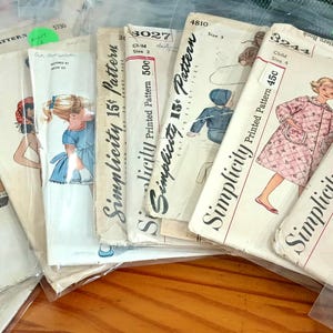 May include: A collection of vintage Simplicity sewing patterns enclosed in clear plastic sleeves. Each paper envelope features black text, numerical identifiers, and colorful illustrations of clothing designs, including dresses and coats for children.