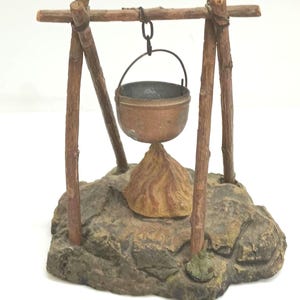 May include: A miniature wooden tripod with a hanging copper pot over a small stone fire pit. The tripod is made of brown twigs and the pot is hanging from a metal chain.