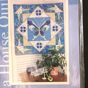 May include: A quilt pattern featuring a butterfly design in shades of blue and white. The quilt has a central butterfly motif, surrounded by geometric patterns and floral accents. The package is labeled "Java House Quilts."