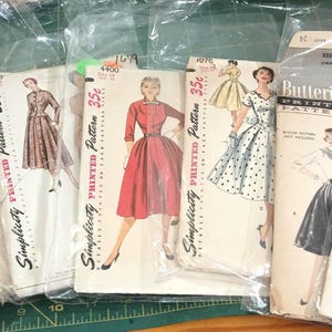 May include: A collection of vintage sewing patterns from Simplicity and Butterick, each enclosed in a clear plastic sleeve. The paper envelopes feature illustrations of various dresses, including styles in red, blue, yellow, and polka dot prints.