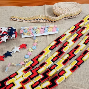 May include: Assortment of decorative ribbons and trims in various designs. Includes red, white, and blue star-shaped trim, floral trim, and zigzag patterns in red, yellow, and black. A roll of beige lace is also present.