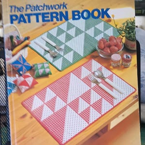 May include: The Patchwork Pattern Book with a red and white placemat, green and white placemat, and small decorative pillows. A bowl of strawberries, small jars, and silverware are also on the wooden table.