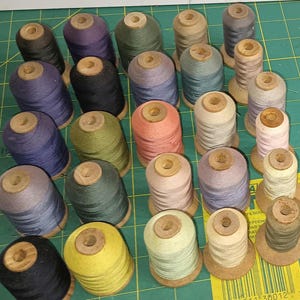May include: A collection of twenty-five spools of thread in various colors, including shades of blue, purple, green, yellow, pink, and neutral tones. Each spool features a wooden core and is arranged in a grid pattern on a green cutting mat.