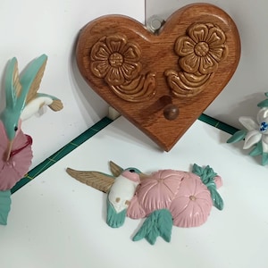 May include: A collection of decorative wall art featuring hummingbirds and floral designs. Includes a wooden heart-shaped wall hook with carved floral details, and several hummingbird and flower sculptures in pink, blue, and white.