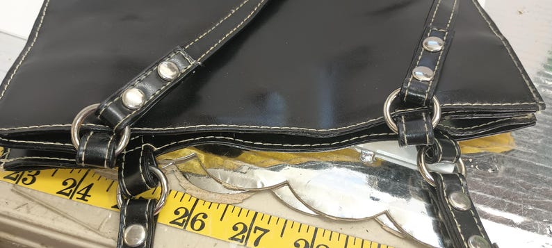 May include: Black tote bag with white stitching and silver hardware. The bag has two straps with silver rivets and metal rings. A yellow measuring tape is visible, showing measurements in inches.