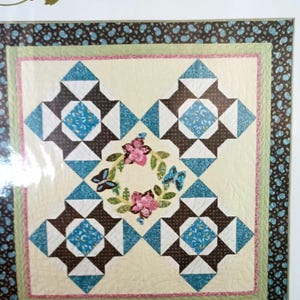 May include: A quilting pattern titled "Quilter's Daydream" from the "A Graceful Stitch" quilt designs by Denise Sheehan. The pattern features a wreath of flowers and butterflies in the center, surrounded by a geometric design of brown, white, and blue squares and triangles.