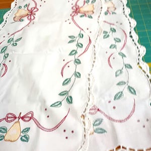 May include: White table runner with a scalloped edge and a crocheted trim. The runner features embroidered pears, green leaves, and red ribbon accents. The design includes small red dots and is suitable for kitchen or dining decor.