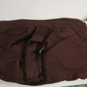 May include: A dark brown clutch purse with a textured fabric. The purse has a gathered design, a black circular handle, and a gold-toned zipper. A tassel hangs from the zipper pull.
