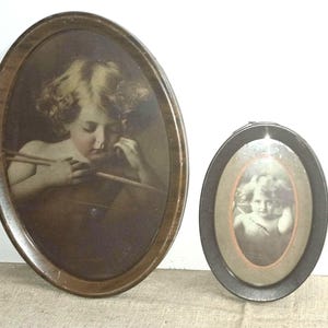 May include: Two oval-shaped wall frames with dark brown wooden borders. Each frame contains a sepia-toned portrait of a young child with curly hair. The larger frame on the left shows the child holding an arrow, while the smaller frame on the right displays a similar portrait of a child with a soft, focused expression.
