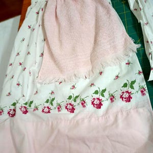 May include: A white apron featuring a small red floral pattern, a wide light pink hem, and a light pink terry cloth towel attached to the front. The apron includes long white fabric ties at the waist and a gathered design at the top section.