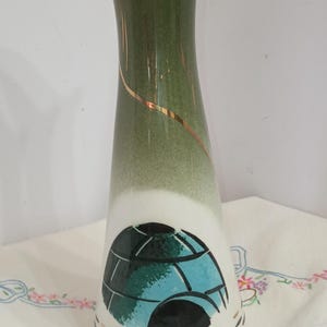 May include: A tall, vintage ceramic vase with a flared top. The vase transitions from a dark green at the top to white at the base, with a gold accent stripe. A blue and black design is featured on the front.