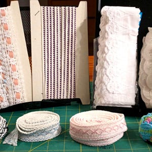 May include: Assortment of decorative ribbons and lace in various colors and styles. Includes white eyelet lace, pink and white patterned lace, and a roll of colorful floral trim. Ideal for sewing and craft projects.