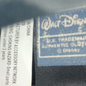 May include: A close-up of a blue Walt Disney label with the text "U.S. TRADEMARK AUTHENTIC CLOTHING". The label is on a black textured fabric. A white label with text is also visible.