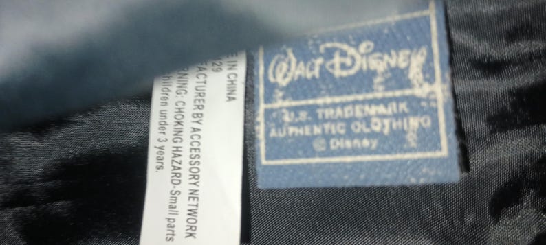May include: A close-up of a blue Walt Disney label with the text "U.S. TRADEMARK AUTHENTIC CLOTHING". The label is on a black textured fabric. A white label with text is also visible.