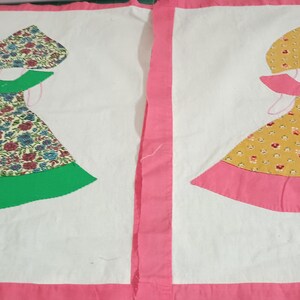 May include: Two handmade fabric panels featuring a girl design. One panel has a floral dress with a green hem and collar, the other has a yellow dress with a pink hem and collar. Both have matching bonnets.