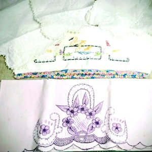 May include: White embroidered linens featuring a swan and floral designs. The top piece has a scalloped edge and a colorful trim. The bottom piece showcases a basket of flowers and swan motifs, all in purple embroidery.