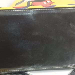 May include: Black rectangular handbag with white stitching and a yellow top featuring a Mickey Mouse design. The bag has a glossy finish and appears to be made of leather or a leather-like material. The bag is a tote style.