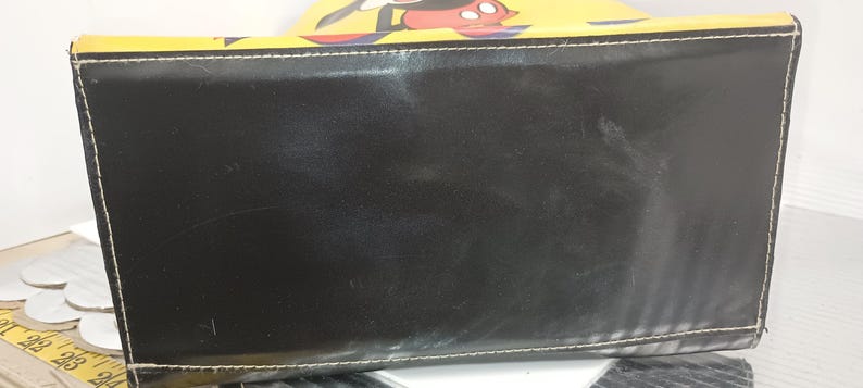 May include: Black rectangular handbag with white stitching and a yellow top featuring a Mickey Mouse design. The bag has a glossy finish and appears to be made of leather or a leather-like material. The bag is a tote style.