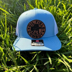 May include: A light blue and white trucker hat with a brown leather patch that says "Oil Field Cowboy" in black lettering. The patch has a black silhouette of a cowboy hat and a pair of boots.