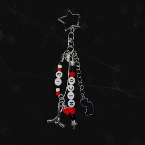 May include: A silver keychain with a star-shaped top and multiple charms. The charms include a hockey stick, the name "NICO" spelled out with white letter beads, red and black beads, and a small outline of a state. The background is black.