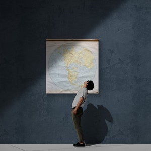 May include: A person stands in front of a world map poster, looking up at the map. The map is a vintage style with a light blue background and yellow continents. The person is wearing a grey shirt and khaki pants.