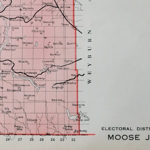 May include: A black and white map of the Moose Jaw electoral district in Saskatchewan, Canada. The map shows the boundaries of the district and the names of the towns and villages within it. The map is titled "ELECTORAL DISTRICT OF MOOSE JAW".