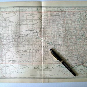 Może przedstawiać: Antyczna mapa Dakoty Południowej, pokazująca granice hrabstw i miasta. Mapa jest otwarta na stronie w oprawionym atlasie, z czarnym długopisem leżącym na stronie.